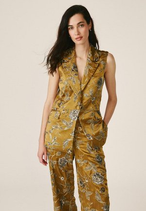 Mujer con cabello largo y oscuro, vestida con un traje sin mangas de color mostaza con bordados florales, de pie con una mano en el bolsillo contra un fondo liso.