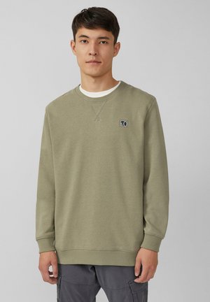 Jeune homme portant un sweat-shirt vert olive clair avec un petit logo en patch sur la poitrine et un pantalon cargo gris foncé, se tenant devant un fond uni.
