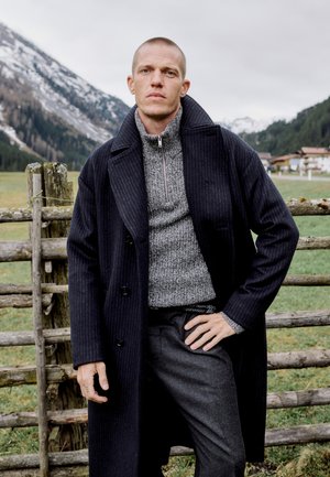 Homme en long manteau sombre et pull gris, debout avec une main sur la hanche près d'une clôture en bois dans un paysage montagneux enneigé.