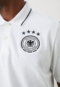 Camisola pólo branca com detalhes em preto, apresentando um logo circular bordado, quatro estrelas em cima e o texto "DEUTSCHER FUSSBALL-BUND". Tecido texturizado.
