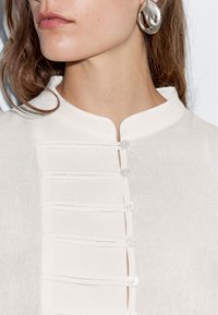 Witte linnenblouse met een opstaande kraag, knoopsluiting met zes knopen, parallelle plooien aan de voorkant en een gestructureerde stof.