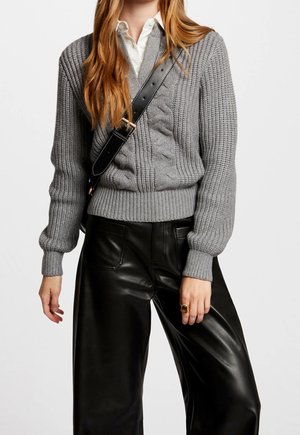 Femme portant un pull en maille torsadée gris, une chemise blanche à col, un pantalon large en cuir noir, un sac bandoulière noir et une bague en or à la main gauche.