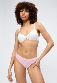 Γυναίκα με σγουρά μαλλιά που φοράει λευκό σουτιέν και ροζ εσώρουχα Tommy Hilfiger, στέκεται μπροστά σε λευκό απλό φόντο.