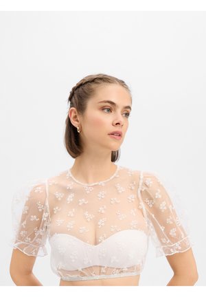 Jeune femme aux cheveux tressés porte un chemisier blanc transparent brodé de fleurs par-dessus un haut blanc sans bretelles, regardant légèrement sur le côté.