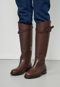 Brown leather knee-high boots with a side zipper, flat sole, and a decorative strap around the top with a metal accent.