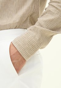 Camicia a righe beige con una texture morbida, caratterizzata da righe bianche. Il polsino è arrotolato, rivelando una tasca bianca dei pantaloni di cotone.