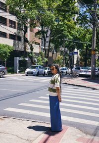 Kvinne med lange fletter og solbriller står ved et fotgjengerfelt i en solrik bygate med trær og parkerte biler.