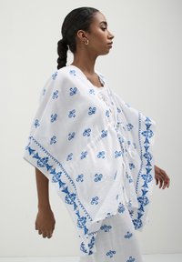 Donna in abito bianco con ricami floreali blu che cammina di lato su uno sfondo bianco uniforme.