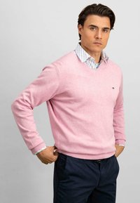 Lys rosa v-hals genser laget av mykt stoff, med ribbestrikkede ermer og hem. Brukt over en blåmønstret skjorte med krage, sammen med mørke bukser.