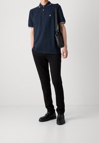 Granatowa koszulka polo z białym wykończeniem kołnierzyka, czarne dopasowane spodnie oraz czarne buty z złotymi dodatkami; modelka trzyma czarną torbę na ramię.