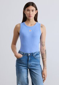 Ανοιχτό μπλε ριμπ αμάνικο τοπ με στρογγυλή λαιμόκοψη και μικρό λογότυπο, σε συνδυασμό με ψηλόμεσο μπλε τζιν παντελόνι.