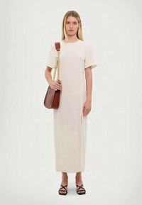 Nais na kreemjas lühikeste varrukatega maxi kleidis, hoides pruuni nahast õlakotti kuldse ketiga rihmaga, jalas mustad vöödiga sandaalid.