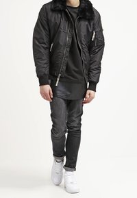 Giubbotto bomber nero con colletto in pelliccia fake, tasche con cerniera, indossato sopra una maglietta nera e abbinato a jeans scuri e sneakers bianche.