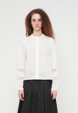 Vrouw met krullend haar, gekleed in een witte blouse met knopen en kantdetails aan de mouwen, en een geplooide donkere rok, staande tegen een effen achtergrond.