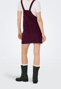 Femme portant une robe salopette en velours côtelé violet par-dessus un haut blanc à manches courtes, avec des chaussettes blanches et des bottes noires mi-mollet, debout de dos.