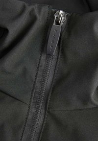 Détail d'une veste zippée en tissu noir, avec un tirant de fermeture éclair gris portant le texte "CORE". Texture lisse avec un design de couture verticale.