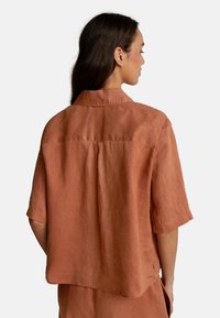 Donna con lunghi capelli scuri che indossa una camicia di lino corta a maniche corte di colore ruggine, vista da dietro su uno sfondo semplice.