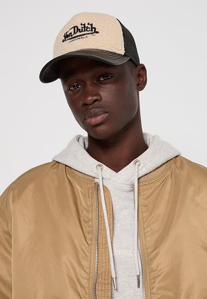 Jeune homme portant une casquette Van Dutch beige et noire, un sweat à capuche gris clair et une veste bomber beige sur un fond gris uni.