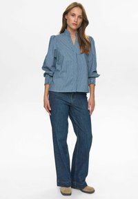 Zila un balta svītrainā blūze ar stāvošu apkakli un pūkainām piedurknēm, kombinēta ar taisnām kājām tumšā denima biksēm un brūniem apaviem.