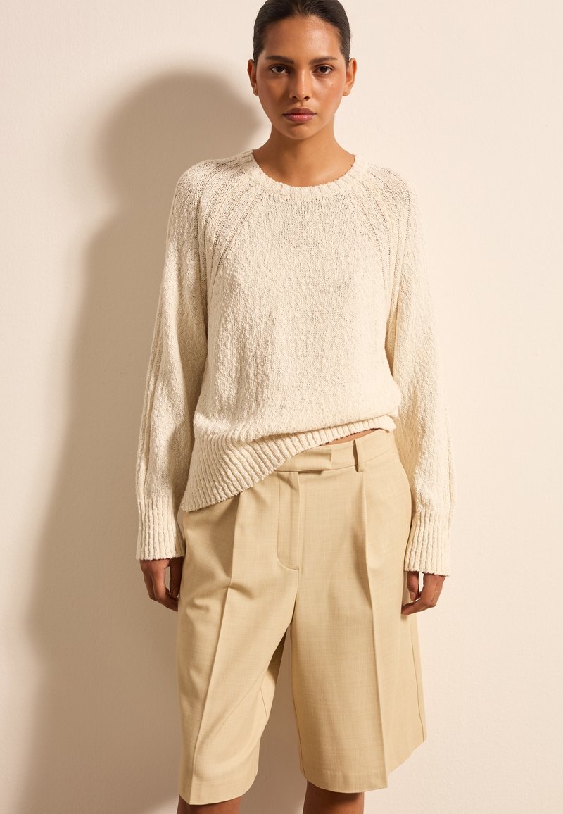 Cremefarbener Strickpullover mit geripptem Saum, kombiniert mit beigen maßgeschneiderten Shorts. Die Shorts verfügen über Falten und eine glatte Textur.