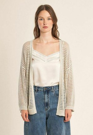 Vrouw draagt een beige opengebreide cardigan, een witte mouwloze blouse met kanten details en een blauw hoogtaille spijkerbroek tegen een effen achtergrond.