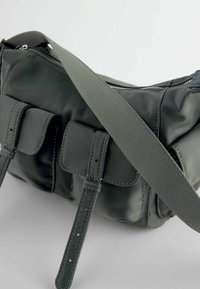 Sac à bandoulière en cuir noir avec deux poches avant à rabat et une sangle en tissu vert foncé réglable sur fond clair.