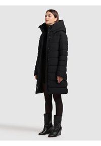 Melns polsterēts garais mētelis ar augstu apkakli, aprīkots ar rāvējslēdzēju un spiedpogas aizdari, un gludu, spīdīgu tekstūru. Valkāts ar melniem potīšu zābakiem.