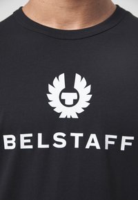 Juodas medvilnės marškinėliai su ryškiu baltu logotipu, turinčiu emblemos virš žodžio "BELSTAFF", apvaliu iškirpimu ir trumpomis rankovėmis.