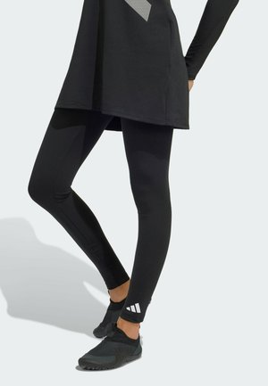 Henkilö, jolla on mustat urheiluleggingsit, joissa on valkoinen Adidas-logo lähellä nilkkaa, ja mustat slip-on tennarit.