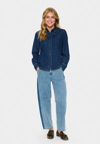 Sieviete smaida un stāv ar rokām kabatās, tērpusies tumši zilā džinsu kreklā, divkrāsu gaišos un tumšos zilos džinsos, kā arī brūnās loafers.