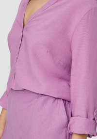 Lichtpaarse linnen blouse met een V-hals, voor knopen en opgerolde mouwen. De stof heeft een zachte textuur met een subtiele glans.