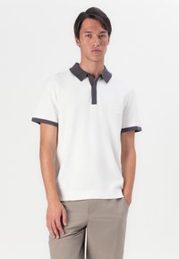 Λευκό μπλουζάκι polo με υφή, σκούρος γκρι γιακάς και μανίκια, με κυκλικό λογότυπο στο στήθος. Εφαρμοστή γραμμή, κοντά μανίκια.