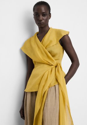 Vrouw draagt een mouwloze gele wikkel-top met gedrapeerde stofdetail over een beige rok, staand tegen een effen witte achtergrond.