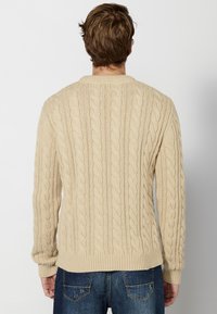 Maglione beige a trecce con polsini e orlo a costine, caratterizzato da un motivo testurizzato e una vestibilità rilassata, abbinato a jeans di denim scuri.