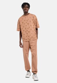 Μπεζ μπλουζάκι σε στυλ πόλο με επαναλαμβανόμενο μοτίβο λογοτύπου, σε συνδυασμό με τζόγκερς σε αντίστοιχο χρώμα που έχουν πλευρικές ρίγες και ελαστικά τελειώματα, φορεμένα με λευκά αθλητικά παπούτσια.