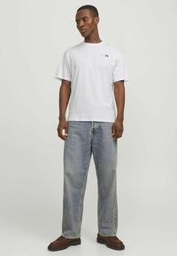 Hvid bomulds T-shirt med korte ærmer og et sort logo på brystet, parret med lyseblå baggy jeans og brune lædersko.