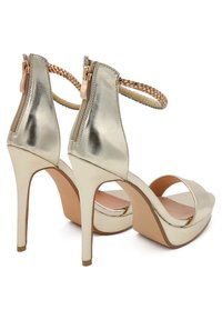Sandali con tacco alto in oro metallico, dotati di una cerniera sul retro, cinturino alla caviglia intrecciato, punta aperta e una suola platform per un'altezza aggiuntiva.
