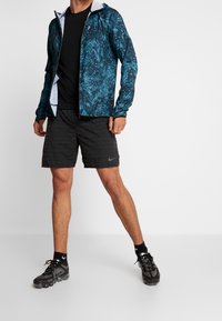 Tenue athlétique comprenant une veste bleue à motifs, un t-shirt noir, un short noir avec logo, et des baskets noires à semelles transparentes avec des chaussettes noires.