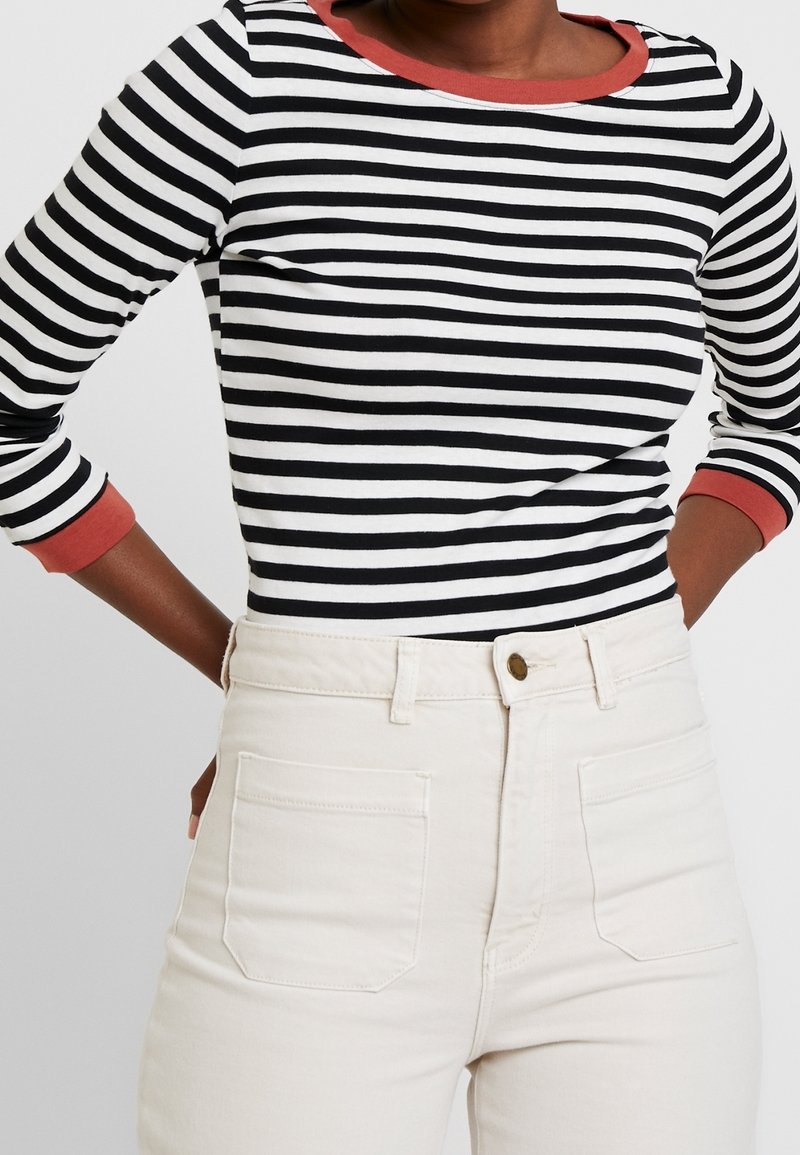 Camicia a maniche lunghe a righe bianche e nere con scollatura rotonda color corallo, abbinata a jeans bianchi a vita alta con tasche posteriori.