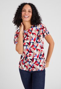 Multikleurige geometrische top van een lichte stof, met korte vlindermouwen en een geplooide halslijn. Gecombineerd met donkere jeans.