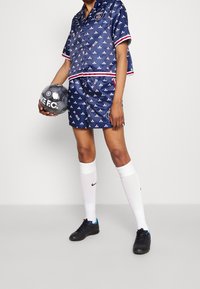 Completo sportivo in satin blu navy con top a fantasia e pantaloni shorts con dettagli bianchi. Calze al ginocchio bianche e scarpe da calcio nere. Tiene in mano un pallone da calcio.