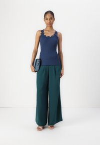 Marineblauw ribbel tanktop met golfachtige randen, gecombineerd met een wijde groene broek en een kleine blauwe handtas. Model draagt lichtgekleurde sandalen.