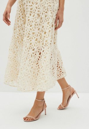 Femme portant une jupe midi en dentelle blanche à fleurs et des sandales à talons hauts roses, marchant sur un sol blanc devant un fond uni.