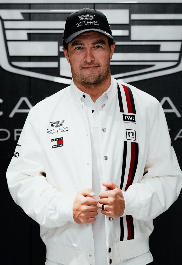 Άνδρας που φοράει λευκό μπουφάν της ομάδας Cadillac Formula 1 και μαύρο καπέλο, στέκεται μπροστά σε μαύρο φόντο με το λογότυπο της Cadillac.