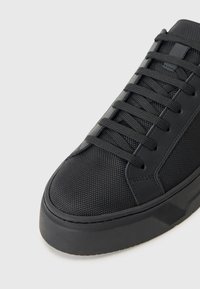 Schwarze Sneaker mit einem strukturierten Mesh-Obermaterial, flachen Schnürsenkeln und einer massiven Gummisohle mit einem subtilen Rillendesign.