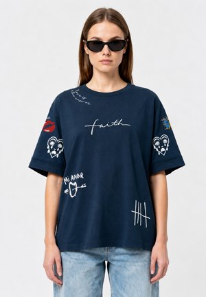 Vrouw met zwarte zonnebril en een marineblauw oversized T-shirt met witte en rode grafische prints en lichtblauwe spijkerbroek.