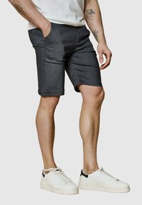 Shorts gris foncé avec poches latérales, coupe slim et tissu légèrement extensible ; portés avec des baskets blanches dotées d'un accent noir.