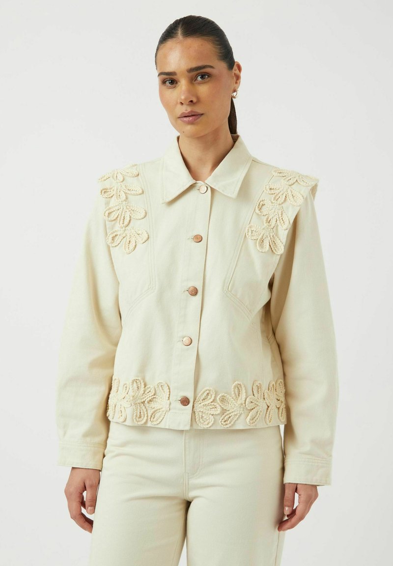 Chaqueta de denim crema con bordados florales en los hombros y en el dobladillo, con cuello clásico y botones de cobre. Textura suave y ajuste relajado.