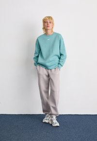 Persona de pie contra una pared blanca, vistiendo un sudadera de color teal, pantalones de chándal gris claro y zapatillas deportivas blancas sobre un suelo de moqueta azul.