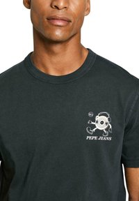 Juodas medvilninis marškinėliai su apvaliu piešiniu, kuriame yra personažas ir tekstas 'Pepe Jeans', su guma apdirbta kaklo iškirpte ir trumpomis rankovėmis.